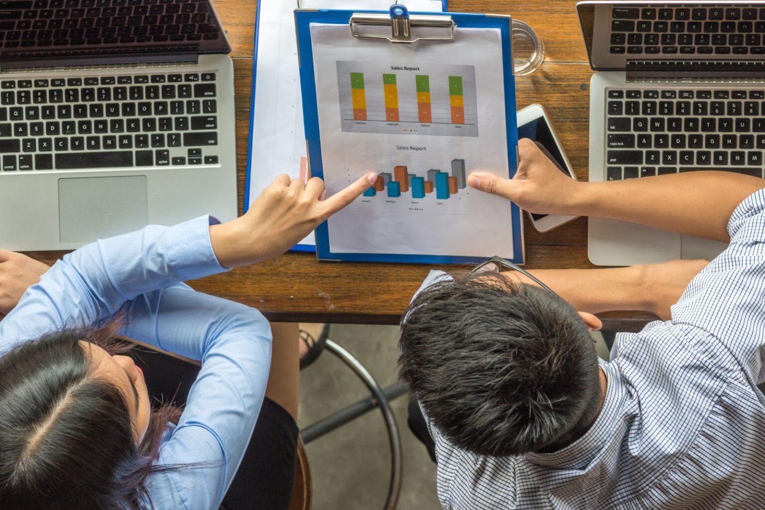 Duas pessoas, vistas de cima, estão sentadas em uma mesa de escritório. Elas analisam um relatório de vendas impresso que contém gráficos de barras. A imagem sugere a discussão de performance de produtos digitais, como a otimização de campanhas de marketing ou o desempenho de aplicativos, com a ajuda de notebooks e celulares.
